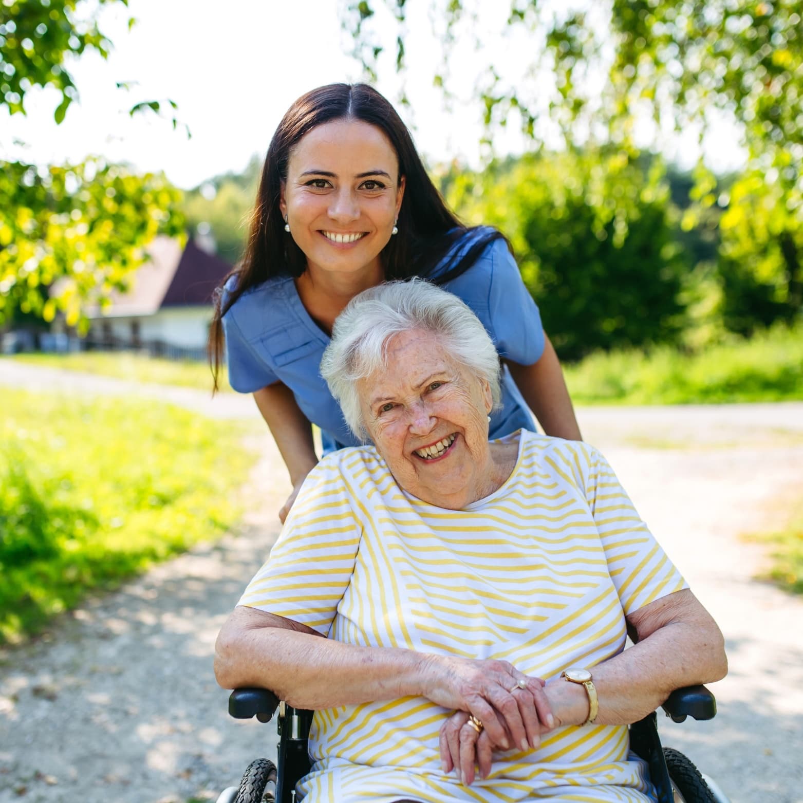 A caregiver in blue scrubs pushing a smiling older woman in a wheelchair outdoors